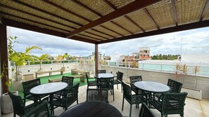 Un espacio de estar al aire libre techado con mesas y sillas, con vistas a una piscina y edificios.