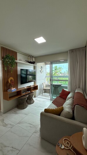 A living room with a TV, sofa, and a balcony with a view. - TNP - Makaranduba 301 (Ipojuca)