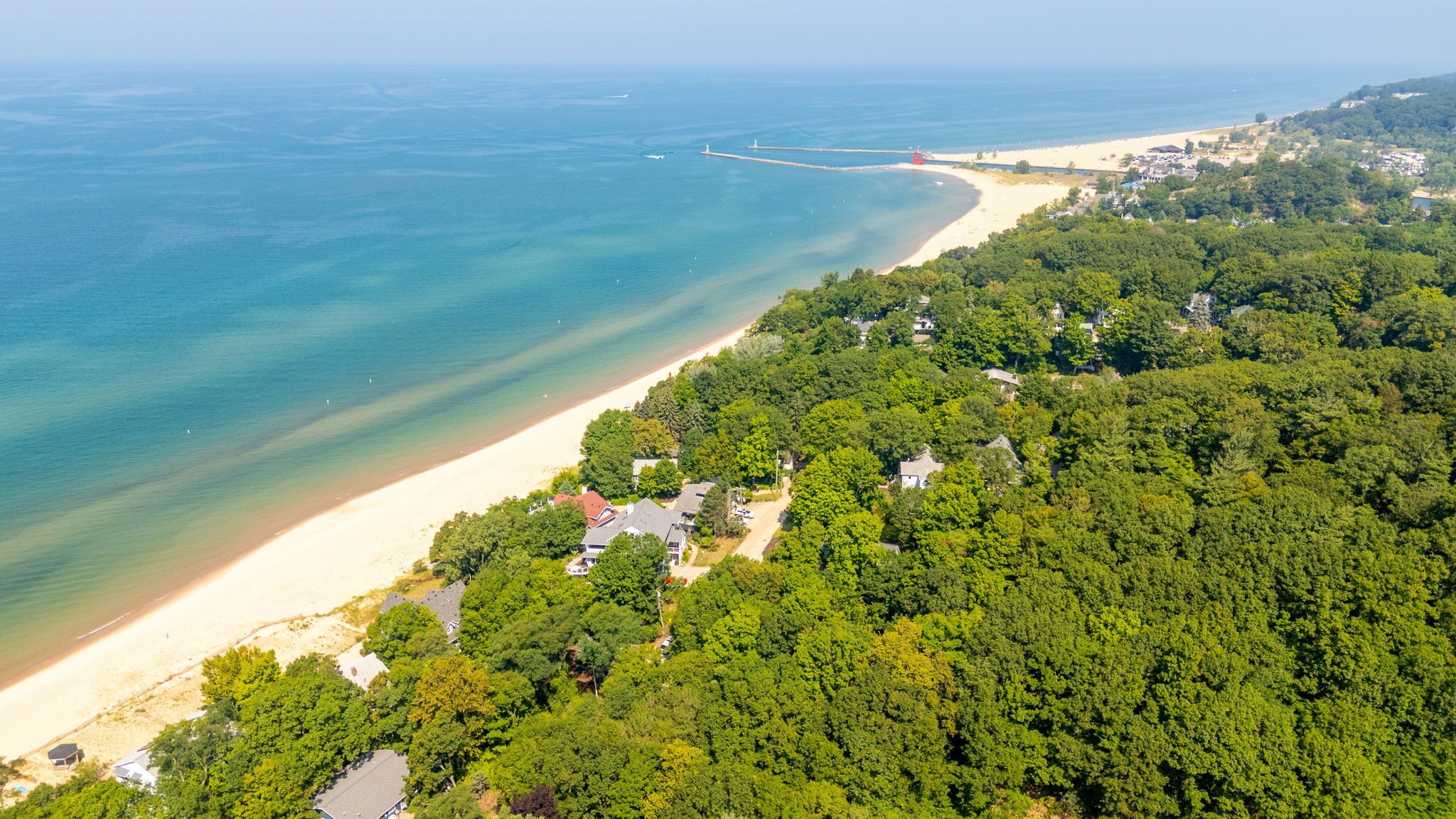 An aerial view of a coastal area with a sandy beach, dense greenery, and scattered buildings.