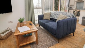 A modern living room with a sofa, a coffee table, a TV, and a kitchen area. - Willow (Barnstaple)