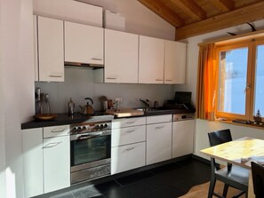 A compact kitchen with white cabinets, a stove, and a dining area with a table and chairs. - Alpatli, Obersaxen-Miraniga, (Obersaxen), 38095B by Interhome (Obersaxen Affeier)