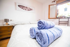 Ein ordentlich bezogenes Bett, gerollte Handtücher, ein Holzkapfenster, eine Nachttischlampe und ein an der Wand hängendes Bild.