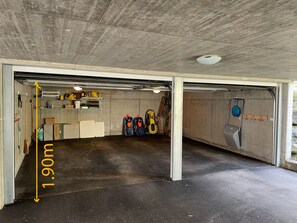 A garage with a concrete floor and walls, containing various items like boxes, a broom, and a blue hanging object. - Casa Carola, Brigels, (Breil/Brigels), 65084B by Interhome (Breil/Brigels)