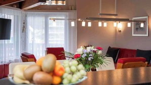 A modern living room with a dining area, featuring a wooden ceiling, a flat-screen TV, a chandelier, and a fruit bowl on the counter. - Arnika Attika, Obersaxen Affeier, (Obersaxen Affeier), 34105B by Interhome (Obersaxen Affeier)
