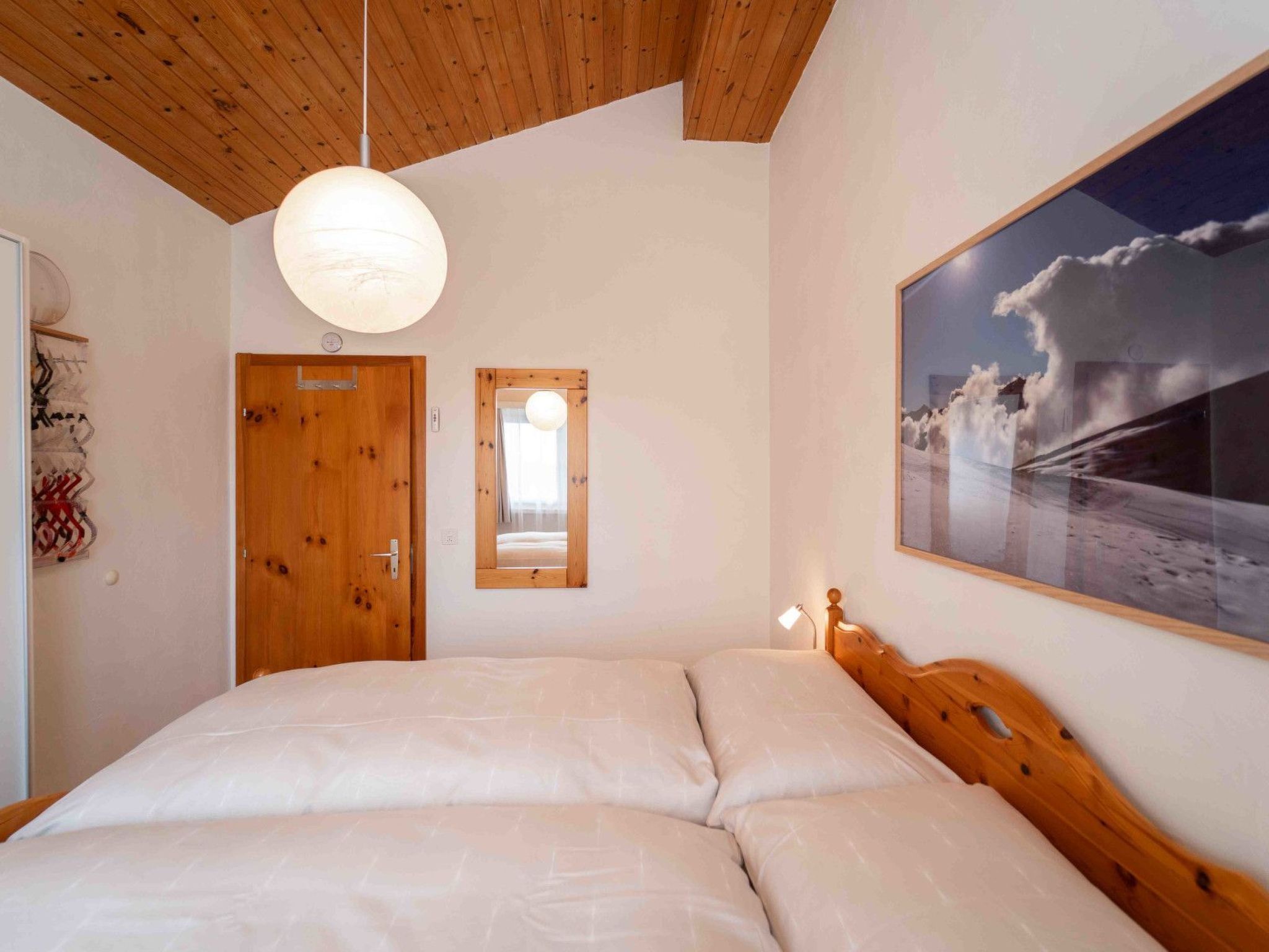 A bedroom with a wooden ceiling, a large bed, a hanging light fixture, a door, a window, and a wall-mounted picture.