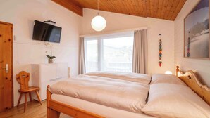 A bedroom with a wooden ceiling, a bed, a TV, a desk, a chair, and a window with curtains. - Arnika Attika, Obersaxen Affeier, (Obersaxen Affeier), 34105B by Interhome (Obersaxen Affeier)