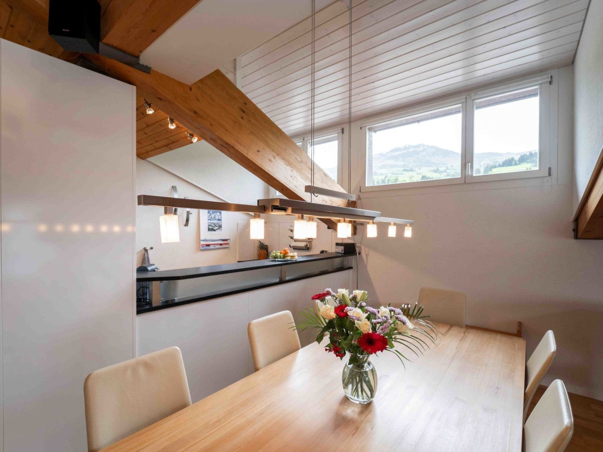 A dining area with a wooden table, chairs, a vase of flowers, and a kitchen in the background.