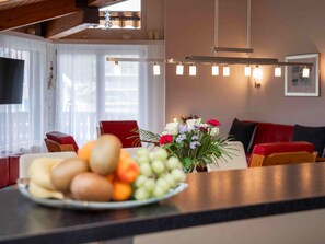 Un salon moderne avec un coin repas, doté d’un plafond en bois, d’un ensemble de canapés, d’une table basse agrémentée de fruits et de fleurs, et d’un téléviseur.