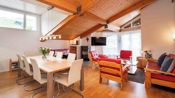 A spacious living area with a wooden ceiling, a dining table with chairs, a red sofa, and a television mounted on the wall. - Arnika Attika, Obersaxen Affeier, (Obersaxen Affeier), 34105B by Interhome (Obersaxen Affeier)