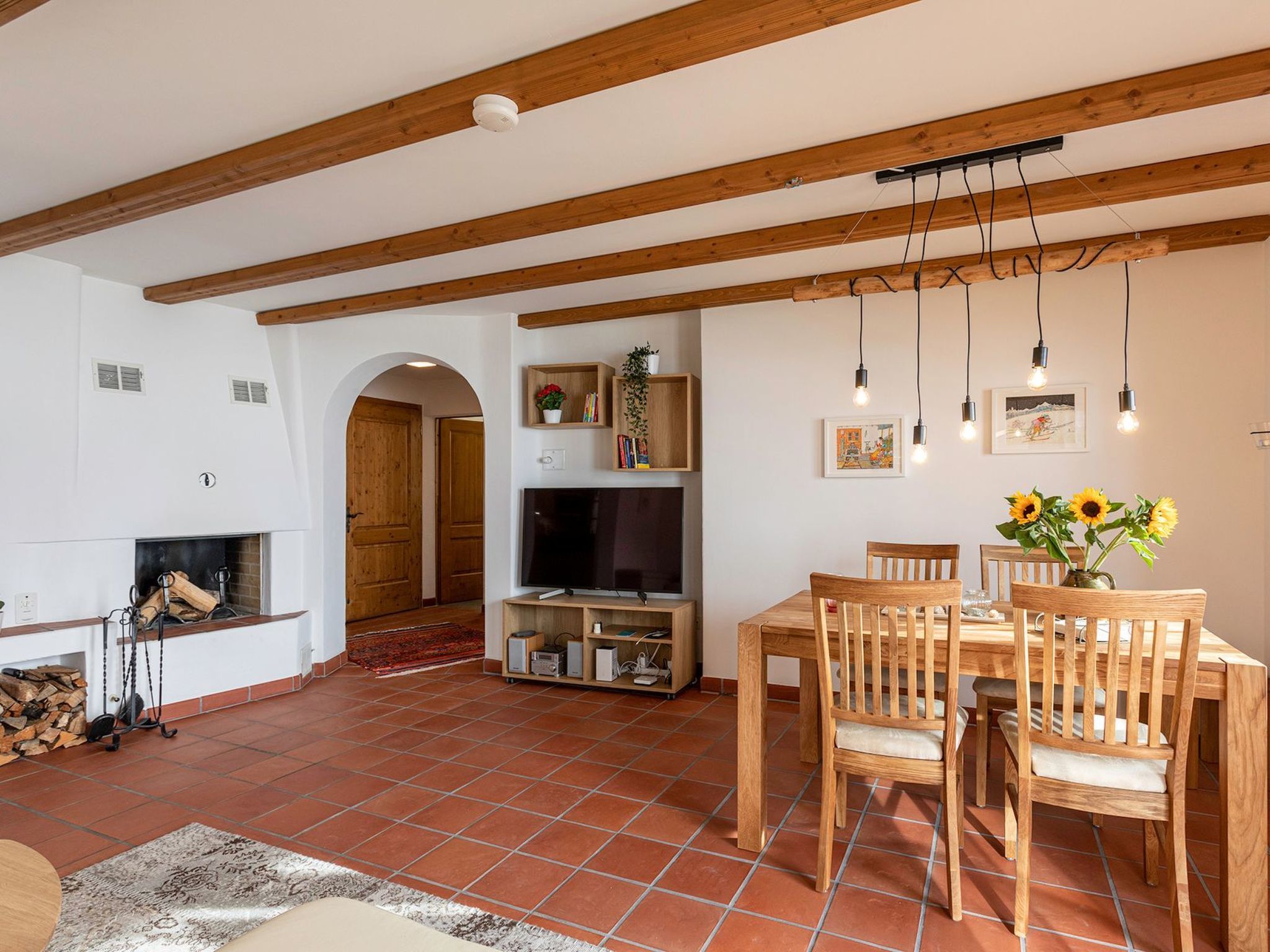 Ein geräumiges Wohnzimmer mit Kamin, Esstisch aus Holz und Stühlen. Es gibt einen Fernseher und Regale mit Büchern und Pflanzen.