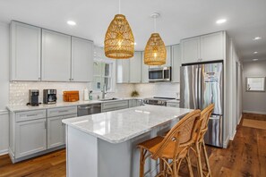 A modern kitchen with white cabinets, a large island, wicker chairs, and pendant lights. - Charming Oceanside Getaway! (Bethany Beach)