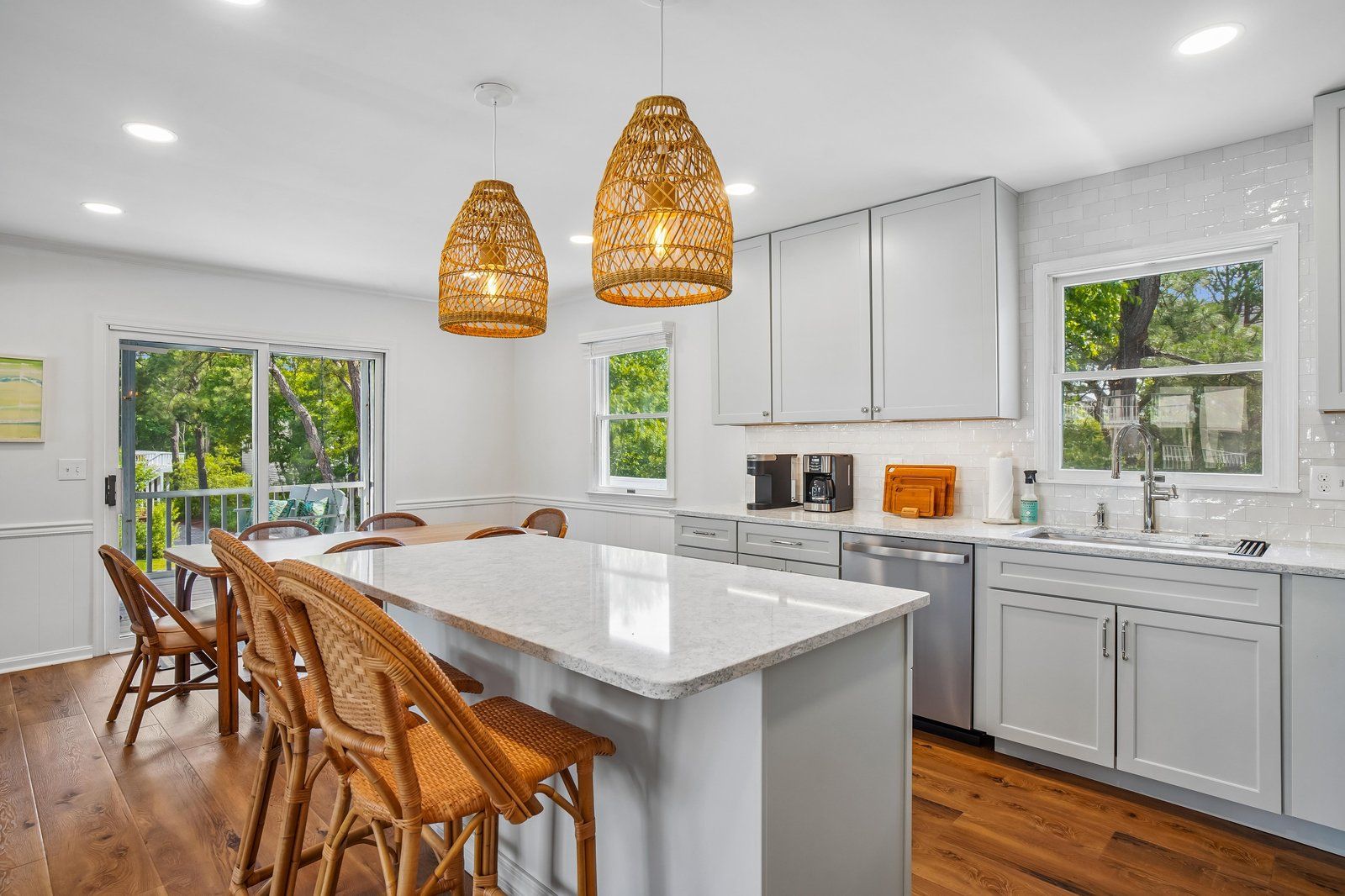 Una cocina moderna con armarios blancos, una isla grande, sillas de mimbre e iluminación colgante.