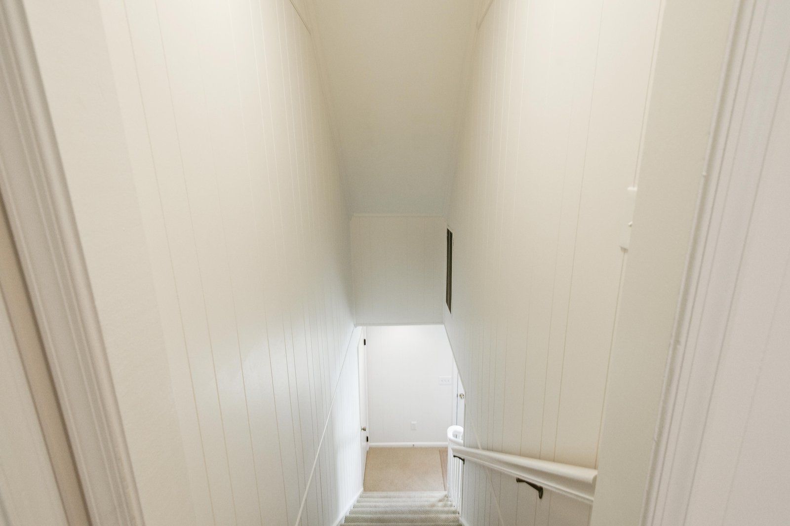 A narrow, white-walled hallway with a carpeted staircase leading to a door at the end.