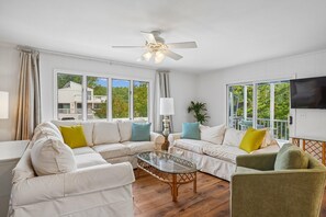 A living room with a sectional sofa, a glass coffee table, a ceiling fan, and large windows with a view of a house and trees. - Charming Oceanside Getaway! (Bethany Beach)