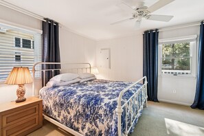 Un dormitorio con cama, mesita de noche, lámpara, ventana con cortinas y ventilador de techo.