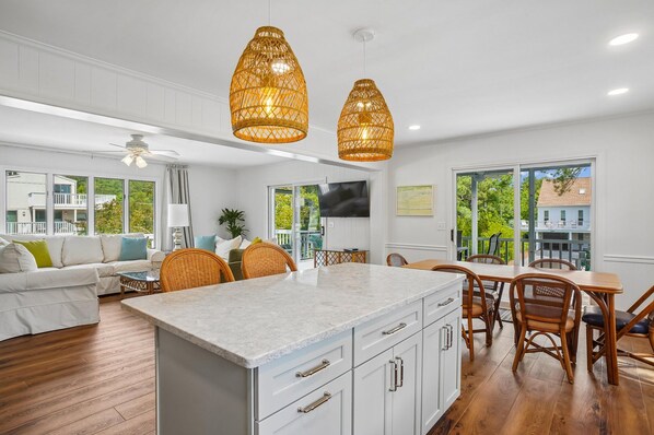 A modern kitchen with a marble island, white cabinetry, and a dining area with wooden chairs and a table. - Charming Oceanside Getaway! (Bethany Beach)