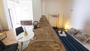 Ein langer Esstisch mit Stühlen, ein kleiner runder Tisch mit Fernbedienung, ein Sofa mit gemusterten Kissen und ein Holzregal mit Kleiderhaken.