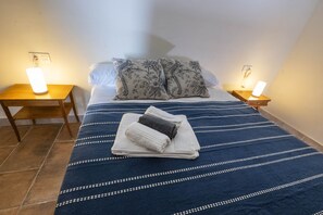 Un lit bien fait, recouvert d’une couverture bleue à rayures, avec deux oreillers assortis et une pile de serviettes pliées. Il y a deux tables de chevet avec des lampes, et la pièce est dotée d’un sol carrelé.