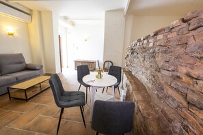 Un salon comprenant un canapé, une table basse et une table à manger avec des chaises. Il y a un mur en pierre et un sol carrelé.