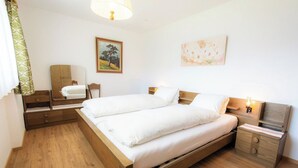 Una cama doble con cabecera de madera, una mesita de noche con lámpara, una silla, un espejo y dos cuadros en la pared.