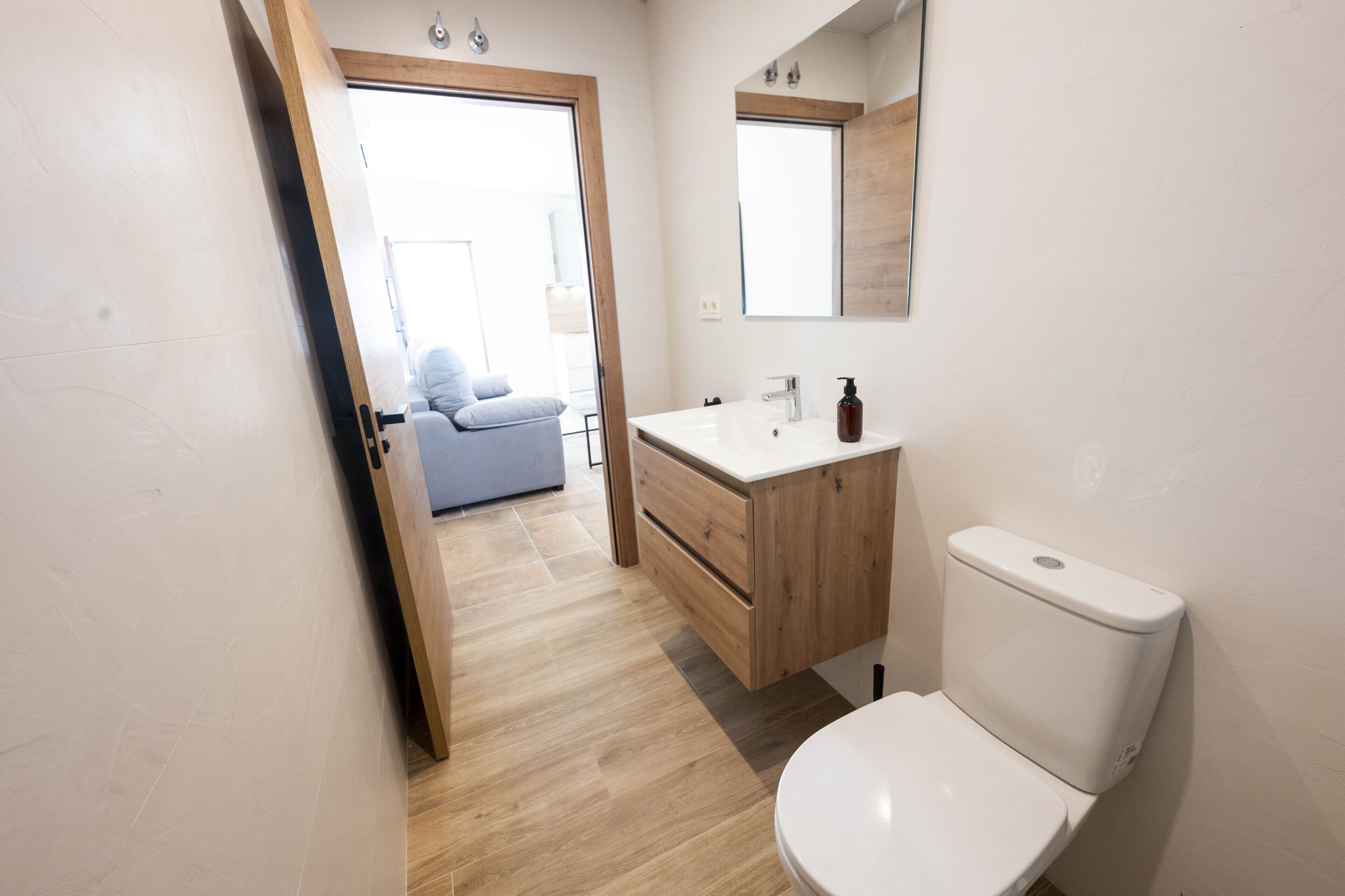 Ein modernes Badezimmer mit Toilette, Waschbecken und Spiegel. Unter dem Waschbecken befindet sich ein Holzkästen und eine Tür zu einem anderen Raum.