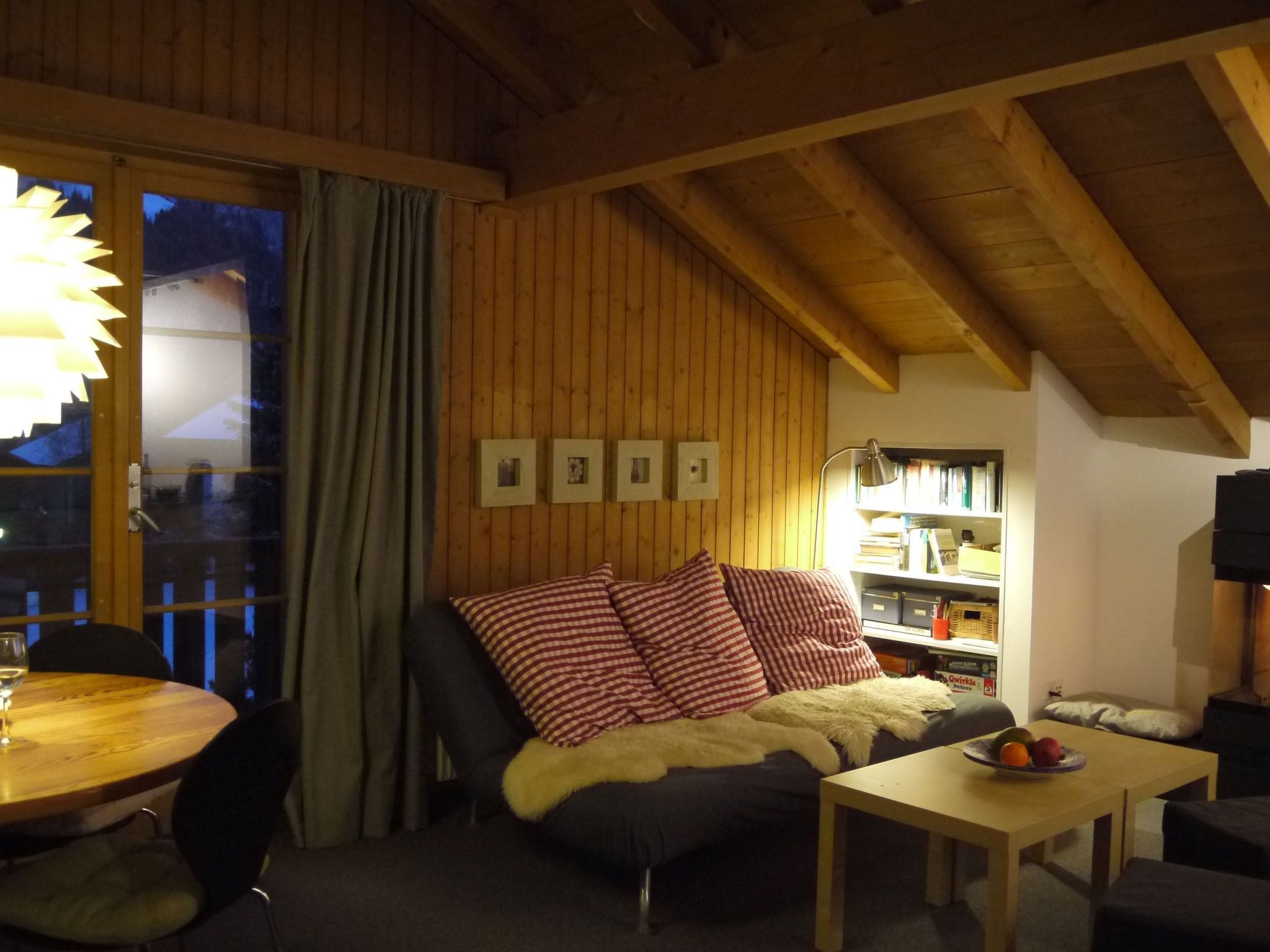 Un salon chaleureux avec un plafond en bois, un canapé orné de coussins à carreaux, une table sur laquelle est posé un panier de fruits et une bibliothèque.