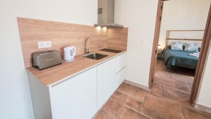 A compact kitchen with a toaster, kettle, and sink, leading to a bedroom with a bed and pillows. - Torre de la Vela Apartment (Granada)