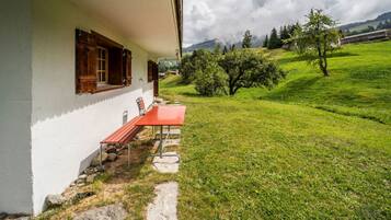 Ein weißes Gebäude mit einer roten Bank davor, einem Steinweg und einem grasbewachsen Feld. Im Hintergrund sind Bäume und Berge zu sehen.