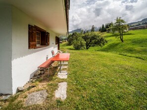 A white building with a red bench outside, a stone pathway, and a grassy field with trees and mountains in the background.