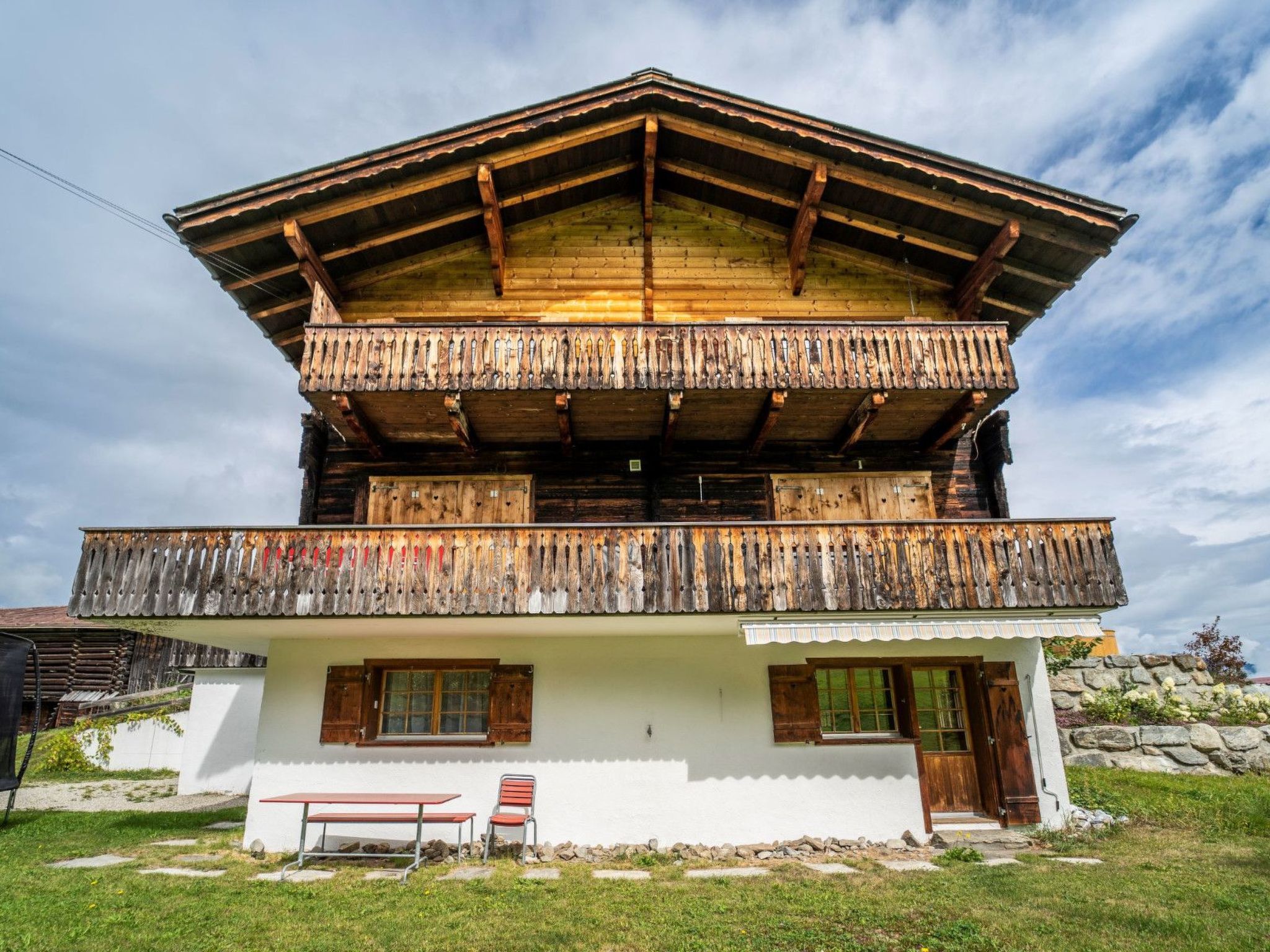 Eine zweistöckige Holzhütte mit Balkon, einer Steinmauer und einer Bank im Vordergrund.