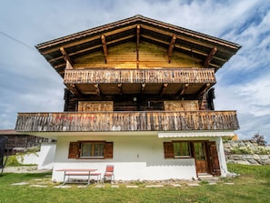 A two-story wooden chalet with a balcony, a stone wall, and a bench in the foreground.