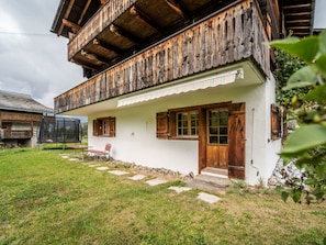 Ein traditionelles Chalet mit Holzbalkon, einem Steinweg, der zum Eingang führt, und einer kleinen Außen-Sitzgelegenheit.