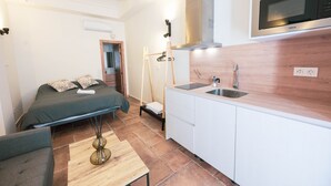 A compact room with a bed, a small sofa, a wooden table, a kitchenette with a microwave, and a sink.