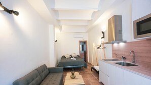 A compact room with a sofa, a small table, a bed, a kitchenette, and a TV mounted on the wall.