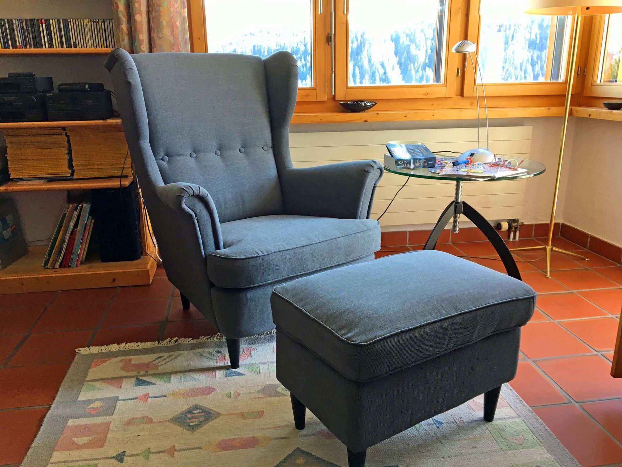 A room with a grey armchair and ottoman, a small glass table with a book and a lamp, and a bookshelf with DVDs and books.