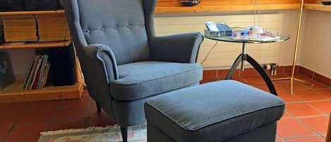 Una habitación con un sillón gris y un reposapiés, una pequeña mesa de vidrio con un libro y una lámpara, y una estantería con DVD y libros.