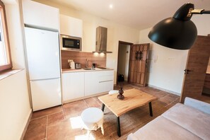 Eine moderne Küche mit weißen Schränken, einer Edelstahl-Mikrowelle und einem eingebauten Backofen. Im Wohnbereich steht ein Holzesstisch mit Lampe, ein kleiner weißer Stuhl und eine Couch.