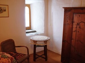 A room with a wooden cabinet, a wicker chair, a small round table with a lace tablecloth, and a doorway leading to another room.