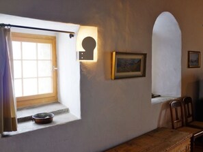 A room with a window, a door, a small window with a view, a painting, and a wooden bench.