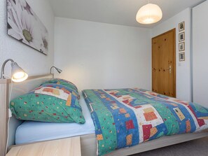 Ein Einzelbett mit einer bunten Steudecke, eine Nachttischlampe und eine Holztür in einem Zimmer mit weißen Wänden.