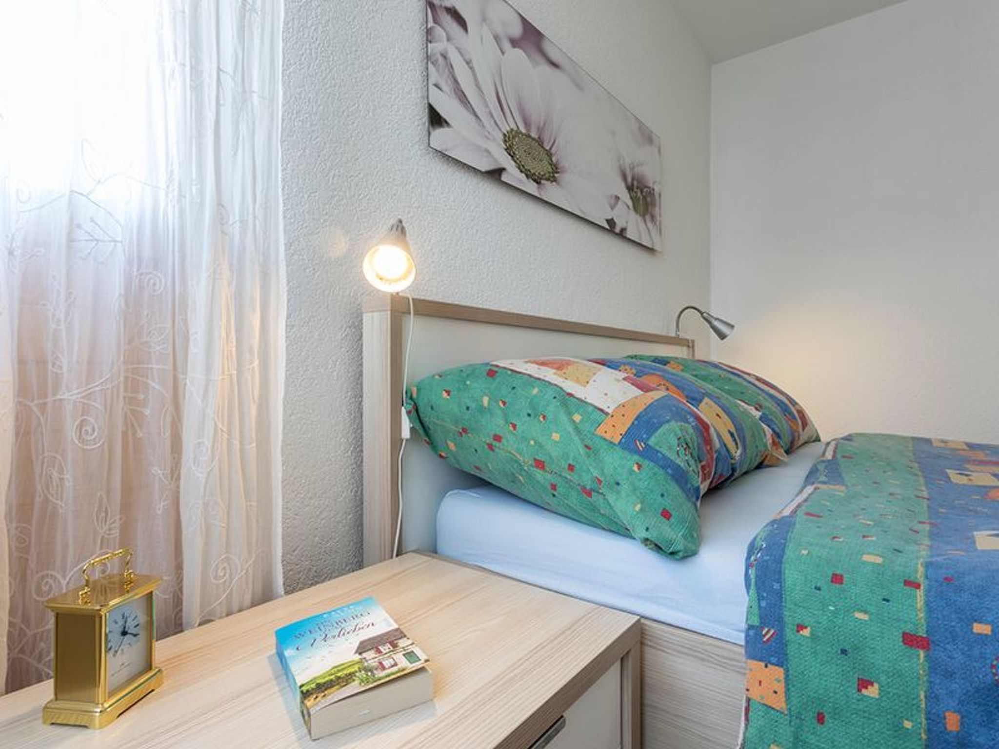 A hotel room with a bed, a bedside table with a book and a clock, a lamp, and a window with curtains.