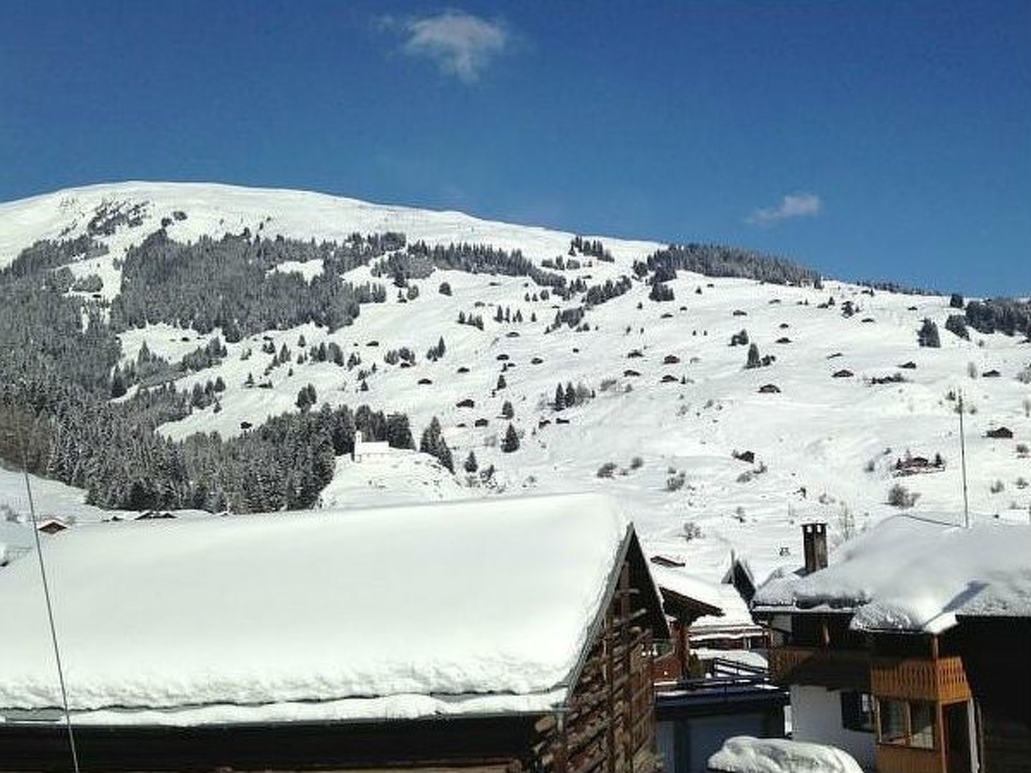 Ein schneegebdeckter Berg mit blauem Himmel. Im Vordergrund liegen Gebäude mit Schneedecken auf den Dächern.