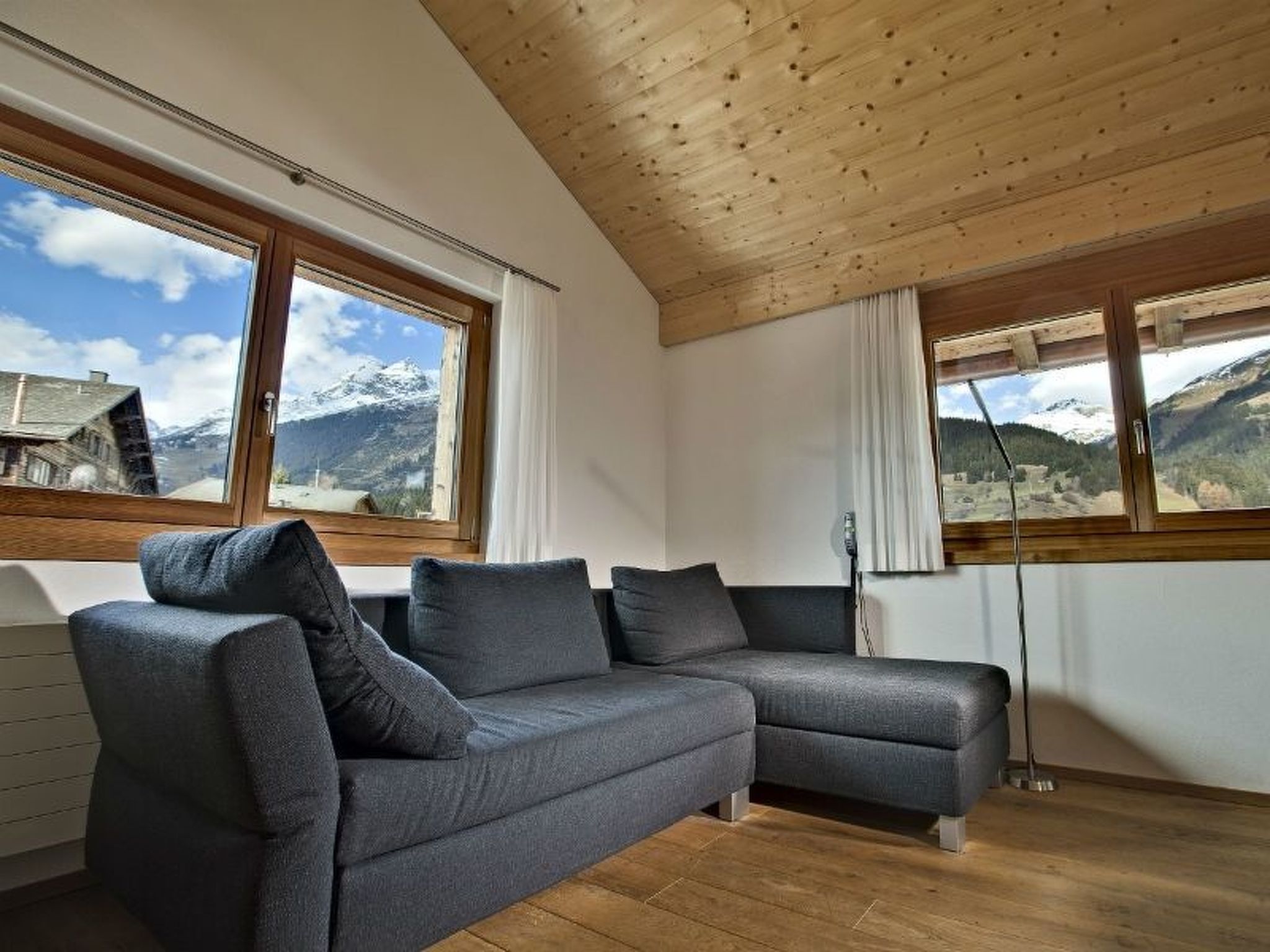Ein modernes Wohnzimmer mit einem grauen Ecksofa, Holzbalkendecke, großen Fenstern mit Bergblick und einer Heizung.
