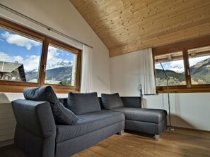 A modern living room with a grey sectional sofa, wooden ceiling beams, large windows with mountain views, and a radiator. - Alpina Schmed, Brigels, (Breil/Brigels), 65002B by Interhome (Breil/Brigels)