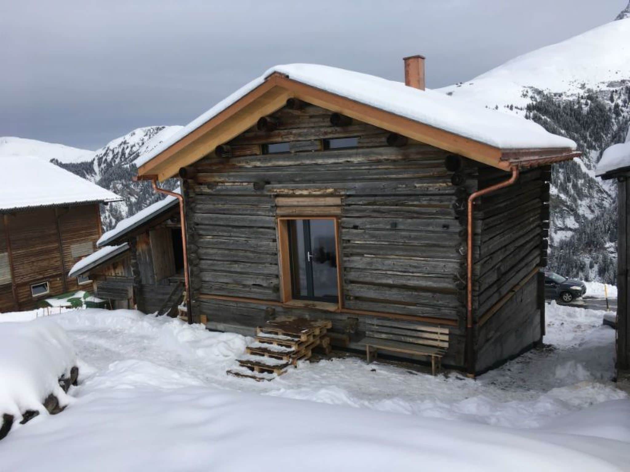 A wooden cabin with a sloped roof and a chimney, surrounded by snow-covered mountains.