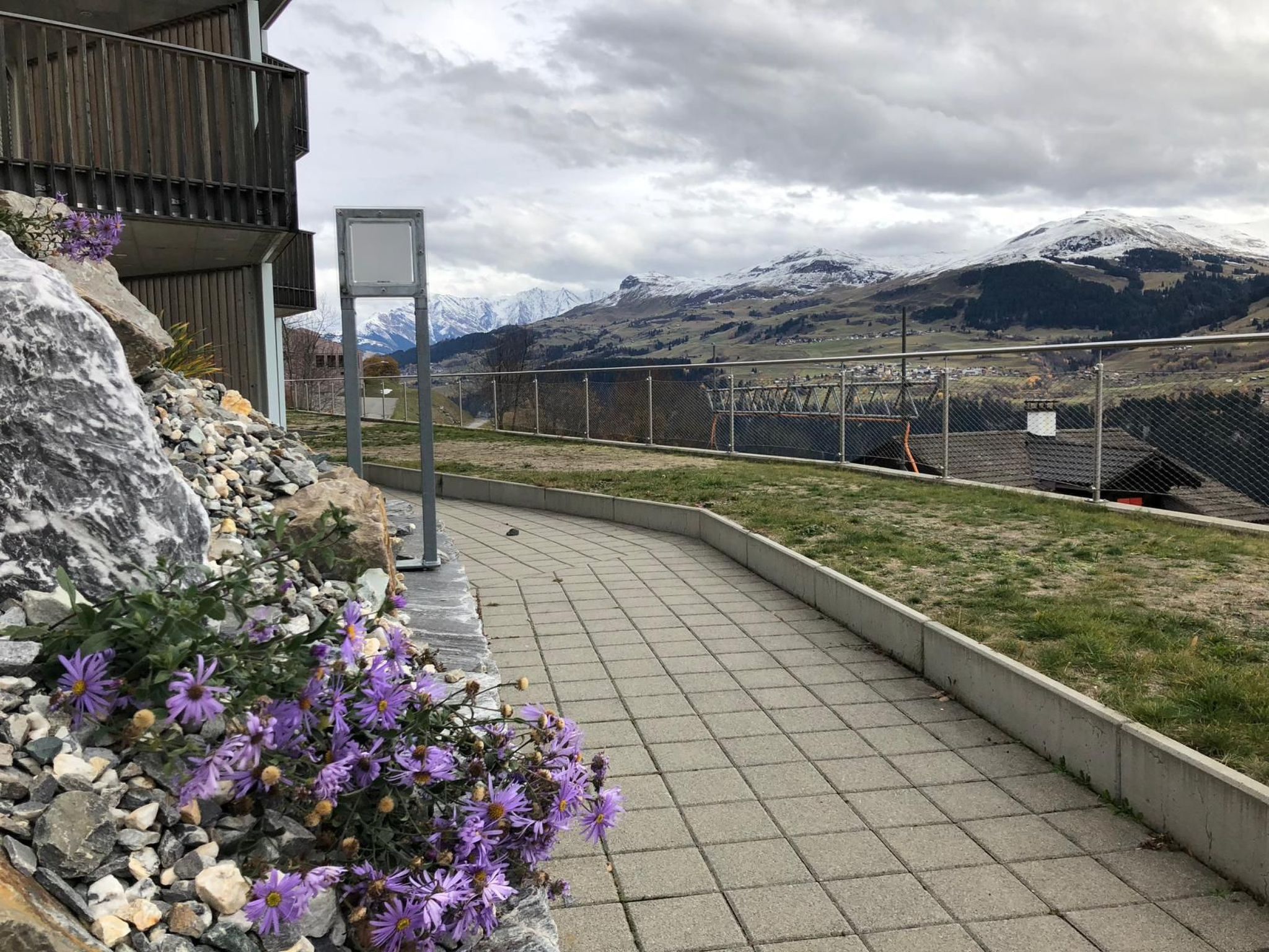 Ein gepflasterter Weg mit Blick auf schneebedeckte Berge, einem eingezäunten Bereich und einem Gebäude mit Holzfassade.