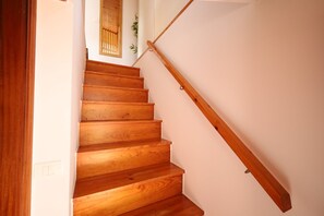 Uma escada de madeira com corrimão, um interruptor de luz na parede e uma janela com persiana de bambu.