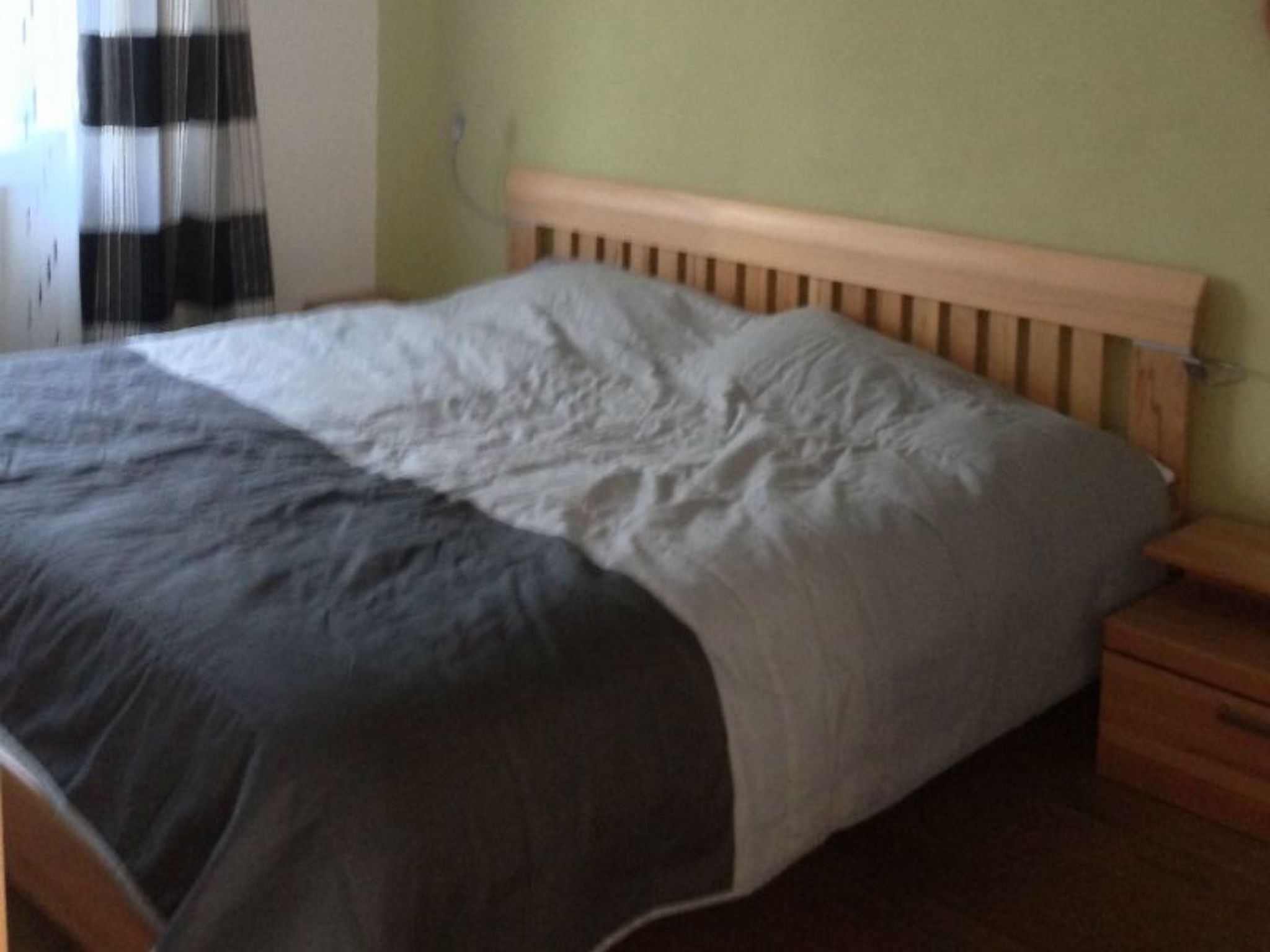 Ein Bett mit Holz-Kopfteil, einer grau-weißen Tagesdecke und einem Nachttisch mit Schublade.