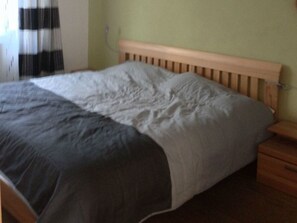 Ein Bett mit Holz-Kopfteil, einer grau-weißen Tagesdecke und einem Nachttisch mit Schublade.