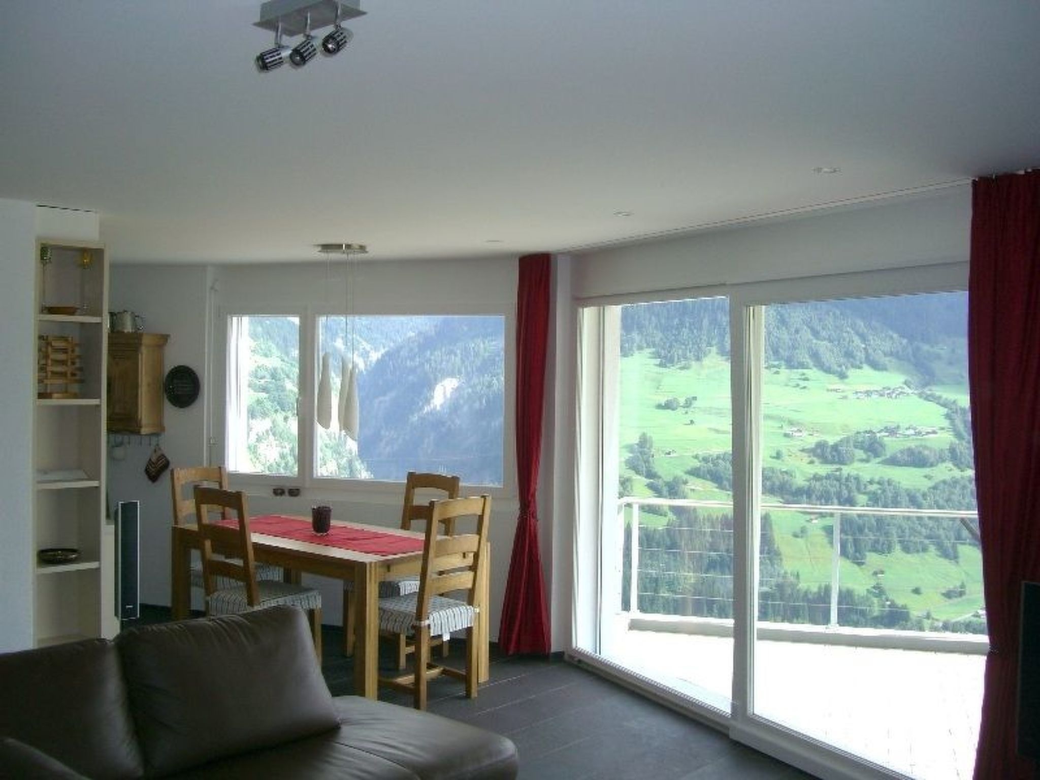 Quarto com mesa de jantar, cadeiras, sofá e grandes janelas com vista para uma paisagem montanhosa.